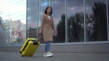 Genç bir kadın sarı bir bavulla yürüyor. Gözlüklü ve ceketli bir kız aynalı bina boyunca yürüyor..