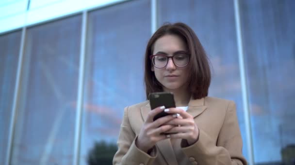 Uma jovem está com um telefone no fundo de um centro de negócios. A garota no casaco está enviando mensagens no smartphone. — Vídeo de Stock