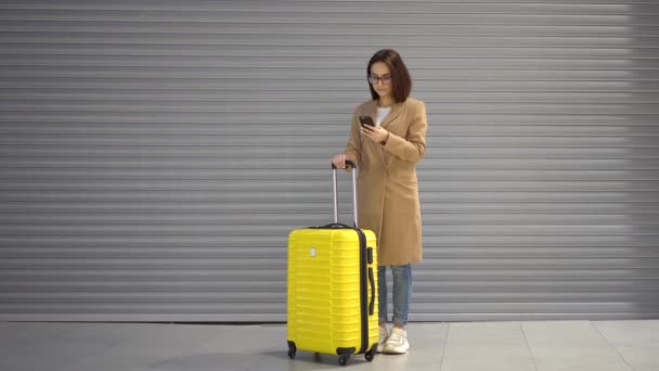 Seorang wanita muda berdiri dengan koper dan telepon di tangannya. Gadis bermantel dengan latar belakang abu-abu. — Stok Video