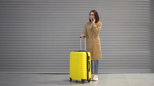 En ung kvinna står med en resväska och pratar i telefon. Flicka i rock på en grå bakgrund. — Stockvideo