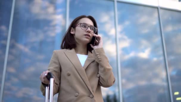 Uma jovem está contra o pano de fundo de um centro de negócios e fala ao telefone. Menina em um casaco com uma mala de fechamento. — Vídeo de Stock