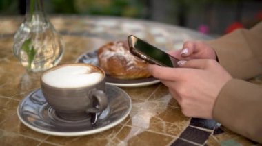 Genç bir iş kadını bir kafede oturuyor ve yakın planda mesajlaşıyor. Kahveli ve çörekli kız yaz verandasında .