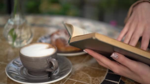 若いビジネスマンの女性がカフェに座って本のクローズアップを読んでいます。夏のベランダでコーヒーとパンを持つ女の子. — ストック動画