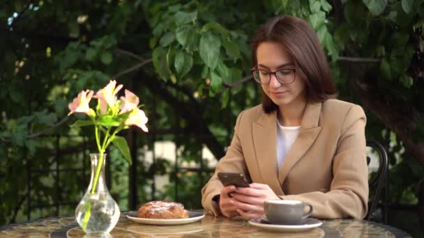 Een jonge zakenvrouw zit in een café en sms 't aan de telefoon. Meisje met koffie en een broodje op de zomerveranda. — Stockvideo