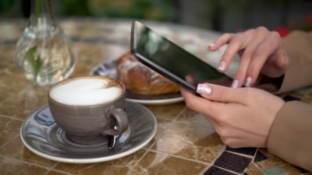 Joven mujer de negocios se sienta en un café y escribe mensajes de texto en una tableta de cerca. Chica con café y un bollo en la terraza de verano. — Vídeos de Stock