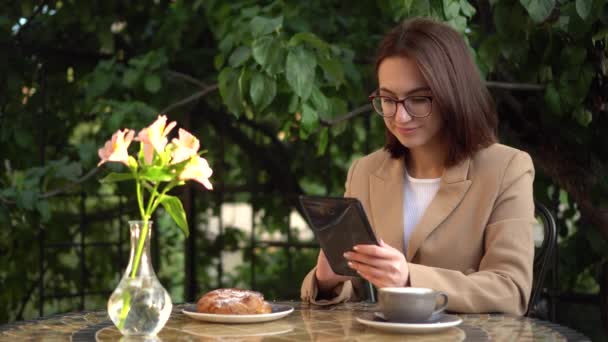 Genç iş kadını bir kafede oturuyor ve bir tablete kısa mesaj yazıyor. Kahveli ve çörekli kız yaz verandasında. — Stok video