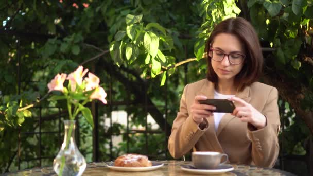 Una giovane donna d'affari si siede in un caffè e fotografa il cibo al telefono. Ragazza con caffè e panino sulla veranda estiva. — Video Stock