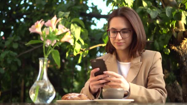 Μια νεαρή επιχειρηματίας κάθεται σε ένα καφέ και στέλνει μηνύματα στο τηλέφωνο. Κορίτσι με καφέ και κουλούρι στην καλοκαιρινή βεράντα. — Αρχείο Βίντεο