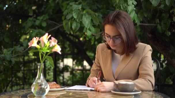 Una joven empresaria se sienta en un café y escribe en un cuaderno. Chica con café y un bollo en la terraza de verano. — Vídeos de Stock