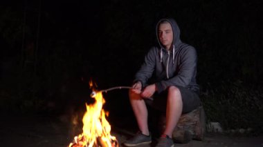 Kapüşonlu genç bir adam ateşin üstünde lokum kızartıyor. Geceleri kamp ateşi.