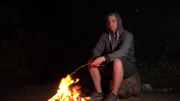 Un giovane col cappuccio frigge i marshmallow su un fuoco e i marshmallow prendono fuoco. Campfire di notte. — Video Stock