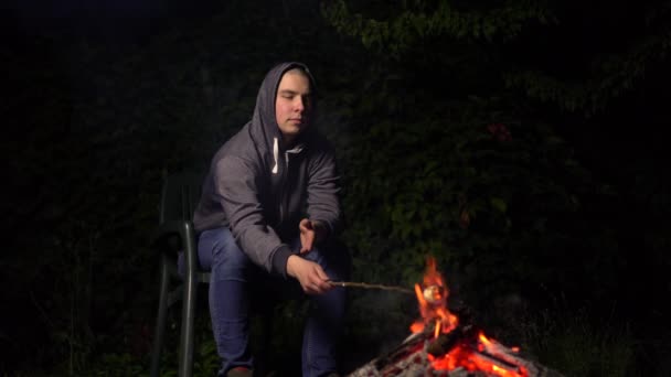 Un jeune homme dans une hotte fait frire des guimauves au-dessus d'un feu et les guimauves prennent feu. Feu de camp la nuit. — Video