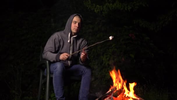 Młody człowiek w kapturze smaży pianki przy ognisku. Ognisko w nocy. — Wideo stockowe