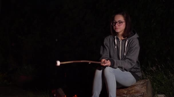 A young woman in a hood fries marshmallows over a fire. Campfire at night. — Stock Video