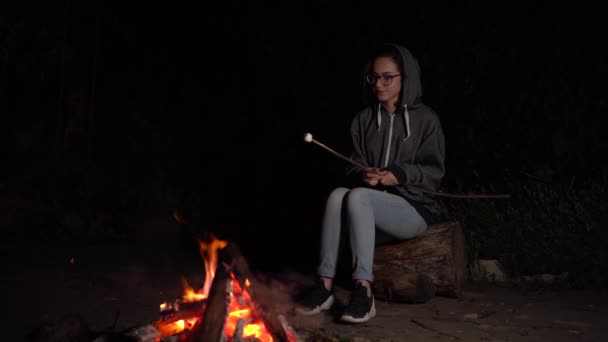 Молодая женщина в капюшоне жарит зефир над огнем. Костер ночью. — стоковое видео