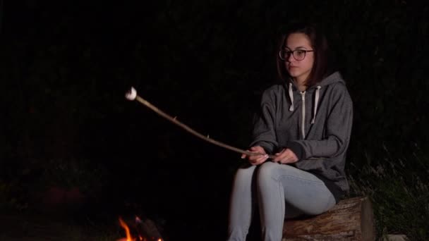 Une jeune femme dans un capot fait frire des guimauves au-dessus d'un feu et les guimauves prennent feu. Feu de camp la nuit. — Video