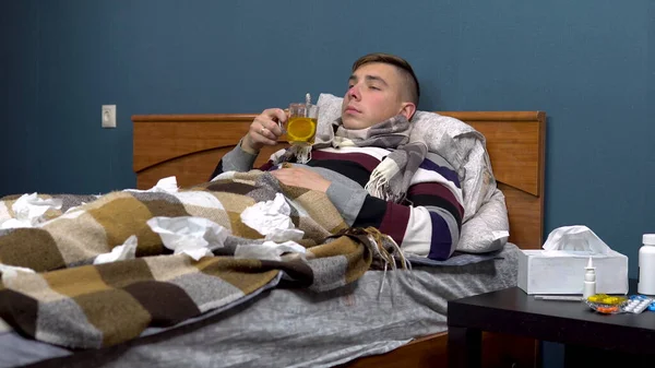 Un joven está bebiendo té con limón. El tipo está enfermo acostado en la cama con una bufanda alrededor del cuello. —  Fotos de Stock