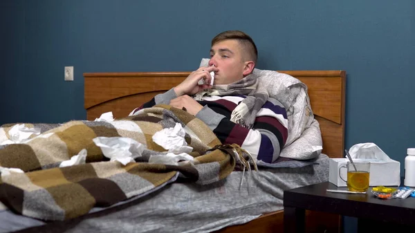 Молодой человек брызгает лекарствами в нос. Парню плохо лежать на кровати с шарфом на шее.. — стоковое фото