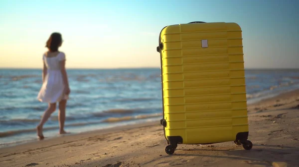 A yellow suitcase stands on the beach against the background of the sea. Travel suitcase. A young woman in a white dress runs in the background. — Stock Photo, Image
