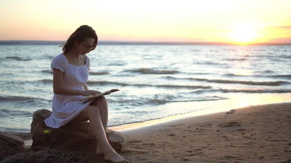 Młoda kobieta siedzi na kamieniu na plaży nad morzem z książką w rękach. Dziewczyna w białej sukience o zachodzie słońca czyta książkę.. — Zdjęcie stockowe