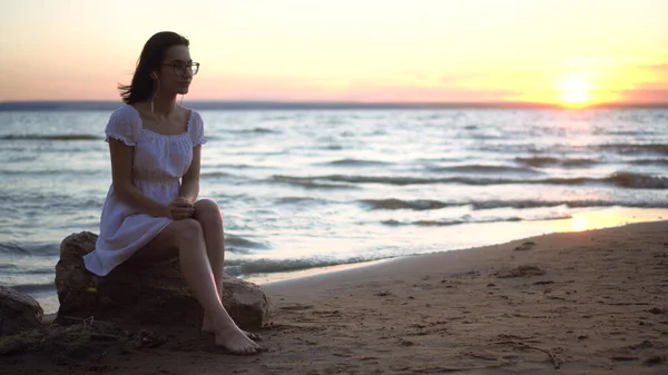 Młoda kobieta siedzi na kamieniu na plaży nad morzem ze słuchawkami w uszach. Dziewczyna w białej sukience o zachodzie słońca słucha muzyki na słuchawkach. — Zdjęcie stockowe