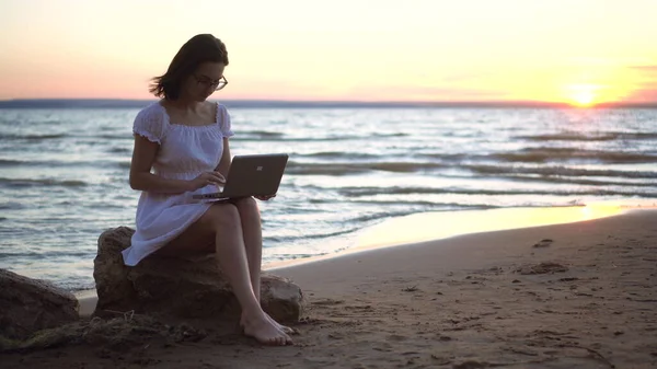 Una mujer joven se sienta en una piedra en la playa junto al mar con un portátil en sus manos. Una chica en un vestido blanco al atardecer está charlando en un portátil. — Foto de Stock