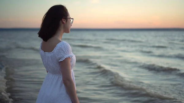 Attraktive junge Frau blickt auf den Sonnenuntergang am Strand am Meer. Das Mädchen im weißen Kleid steht mit dem Rücken. — Stockfoto