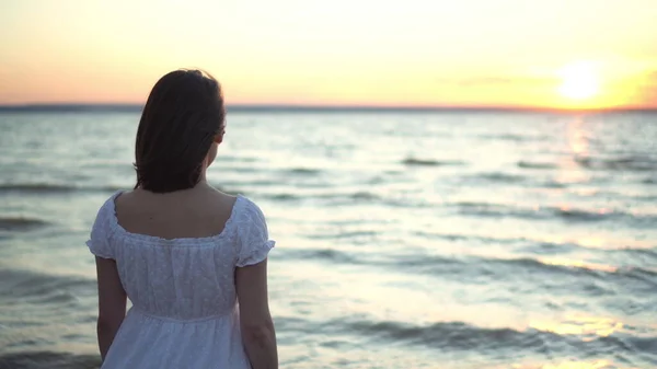 Attraktive junge Frau blickt auf den Sonnenuntergang am Strand am Meer. Das Mädchen im weißen Kleid steht mit dem Rücken. — Stockfoto