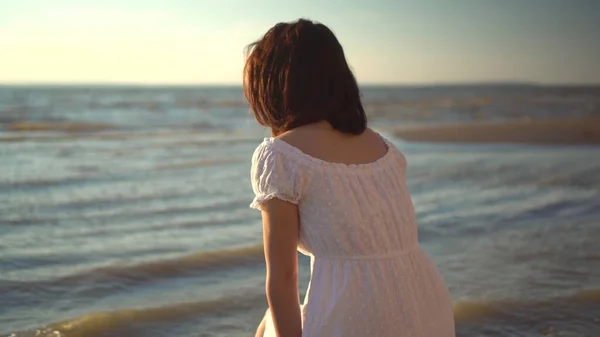 Çekici genç bir kadın sahilde oturur ve denize bakar. Beyaz elbiseli kız sırt üstü oturuyor.. — Stok fotoğraf