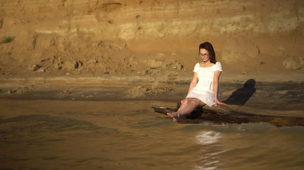 Uma jovem atraente está sentada num tronco junto ao rio. Uma menina em um vestido branco espirra as pernas no rio contra o fundo de uma colina arenosa. — Fotografia de Stock