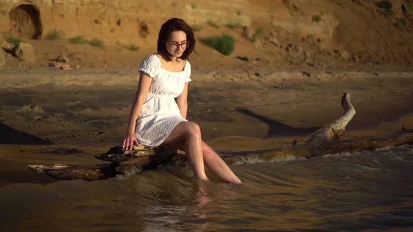 Uma jovem atraente está sentada num tronco junto ao rio. Uma menina em um vestido branco espirra as pernas no rio contra o fundo de uma colina arenosa. — Fotografia de Stock