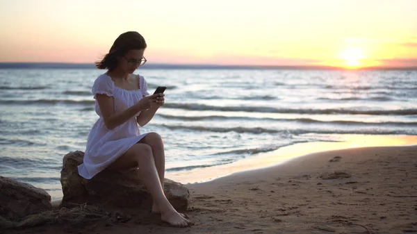 Genç bir kadın sahilde bir taşın üzerinde oturuyor. Elinde telefon var. Gün batımında beyaz elbiseli bir kız. — Stok fotoğraf