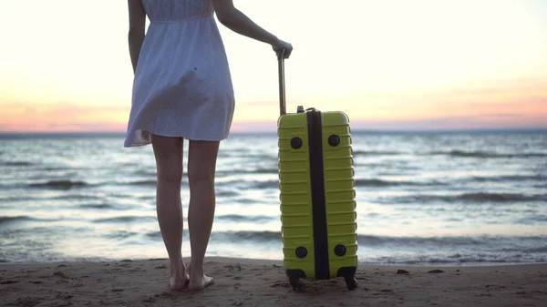 Junge Frau mit gelbem Koffer am Strand am Meer. Ein Mädchen im weißen Kleid betrachtet den Sonnenuntergang aus nächster Nähe. — Stockfoto