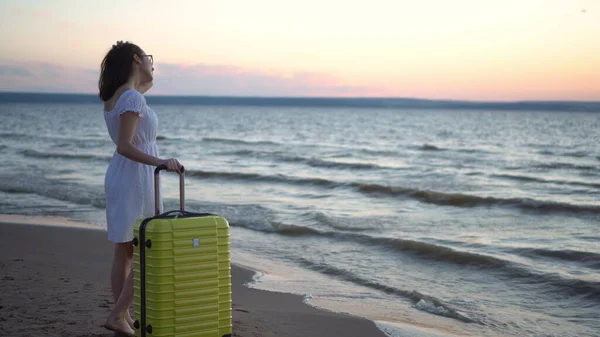 Mujer joven con una maleta amarilla en la playa junto al mar. Una chica con un vestido blanco mira el atardecer. — Foto de Stock