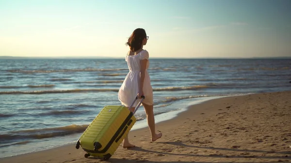 Mujer joven camina por la playa del mar con una maleta amarilla. Una chica vestida de blanco camina descalza sobre la arena. — Foto de Stock