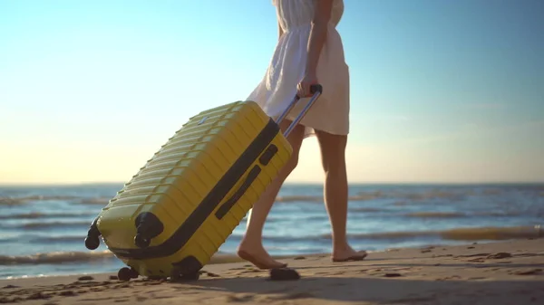Giovane donna cammina lungo la spiaggia del mare con una valigia gialla primo piano. Una ragazza con un vestito bianco cammina scalza sulla sabbia. — Foto Stock
