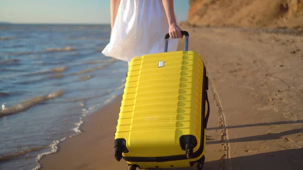 Young woman walks along the beach of the sea with a yellow suitcase closeup. A girl in a white dress walks barefoot on the sand. — Stock Photo, Image