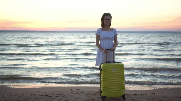 Young woman with a yellow suitcase on the beach by the sea. Girl in a white dress by the sea at sunset. — Stock Photo, Image