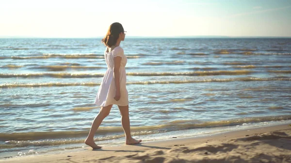 Atrakcyjna młoda kobieta spaceruje wzdłuż plaży morskiej. Dziewczyna w białej sukience spaceruje boso po plaży. — Zdjęcie stockowe