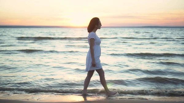 Atraktivní mladá žena kráčí po pláži při západu slunce. Dívka v bílých šatech kráčí bosá po pláži. — Stock fotografie