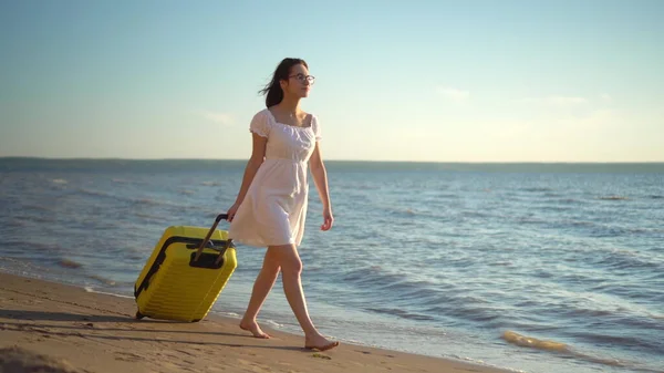 Giovane donna cammina lungo la spiaggia del mare con una valigia gialla. Una ragazza con un vestito bianco cammina scalza sulla sabbia. Viaggia attraverso le fatiche. — Foto Stock