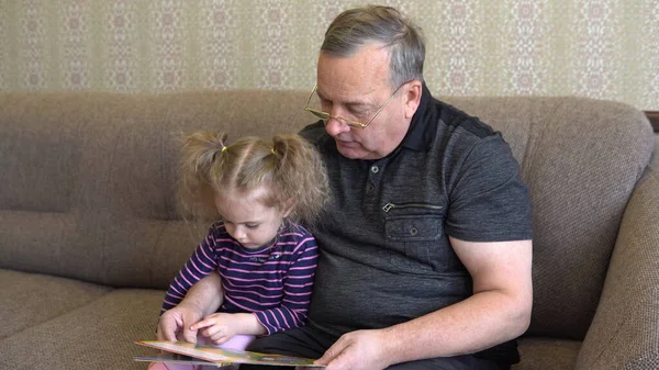 A neta está a ler um livro com o avô. A menina franze a testa no livro e ouve atentamente o avô. Sentados no sofá juntos close-up — Fotografia de Stock