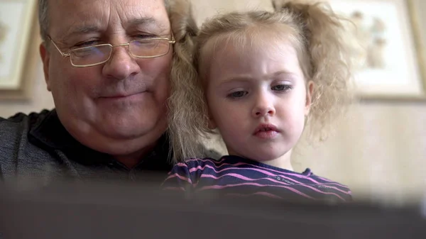Petite-fille et grand-père sont assis avec une tablette. La fille fronce les sourcils à la tablette. Assis sur le canapé ensemble gros plan. — Photo