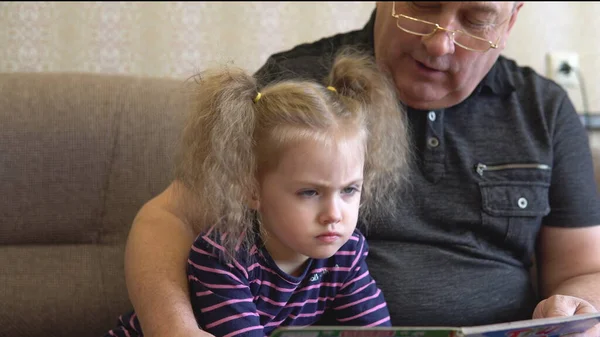Sa petite-fille lit un livre avec son grand-père. La fille fronce les sourcils au livre et écoute attentivement Grand-père. Assis sur le canapé ensemble gros plan — Photo