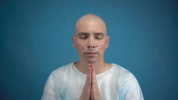 Bald young man with oncology looks at the camera and prays on a blue background. The patient folded his hands and prays with a smile. Hair loss due to chemotherapy.