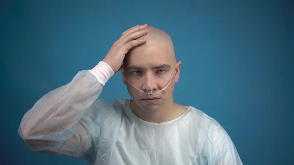 Un giovane calvo con l'oncologia guarda tristemente la macchina fotografica su uno sfondo blu. Il paziente tocca la sua testa calva. Perdita di capelli a causa della chemioterapia. — Foto Stock