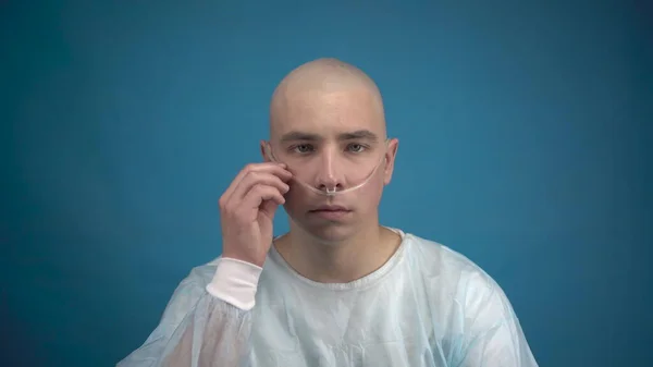 Egy kopasz fiatalember onkológiával szomorúan néz a kamerába kék háttérrel. A beteg kiegyenesíti a légzőkészülék csövét. Kemoterápia miatti hajhullás. — Stock Fotó