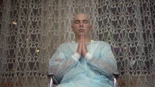 Egy kopasz, rákos fiatalember a kamerába néz és imádkozik. A beteg összecsukta a karját és tolószékben imádkozott otthon. Kemoterápia miatti hajhullás. — Stock Fotó