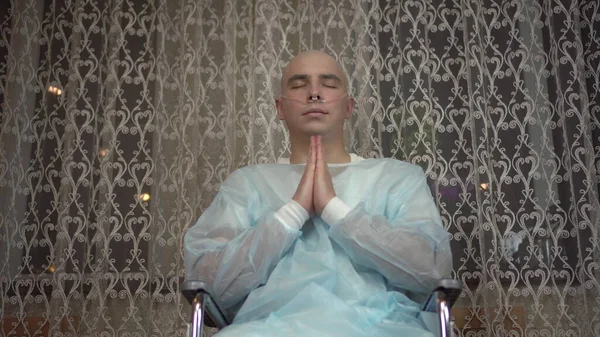 En skallig ung man med cancer tittar på kameran och ber. Patienten vek armarna och ber med ett leende hemma i rullstol. Håravfall på grund av kemoterapi. — Stockfoto