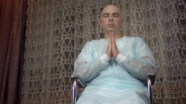 A bald young man with cancer looks at the camera and prays. The patient folded his arms and prayed in a wheelchair at home. Hair loss due to chemotherapy.